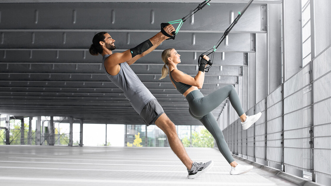 En mand og en kvinde træner i en underjordisk parkeringskælder med et TRX-bånd